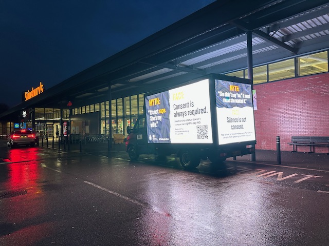 A Transit Media digital advertising van outside a Sainsbury's at night, with brightly lit campaign panels visible on both sides displaying Merseyside Police serious sexual offences awareness messaging including 'Consent is always required' and 'Silence is not consent'